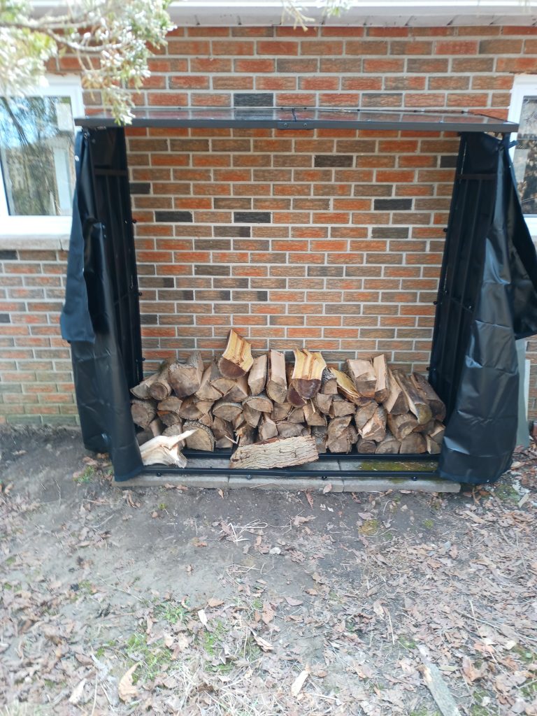 A large wood storage rack about 2m wide and high, and about 600mm deep, set on concrete tiles and against a brick wall. Black covers hang open at each end.