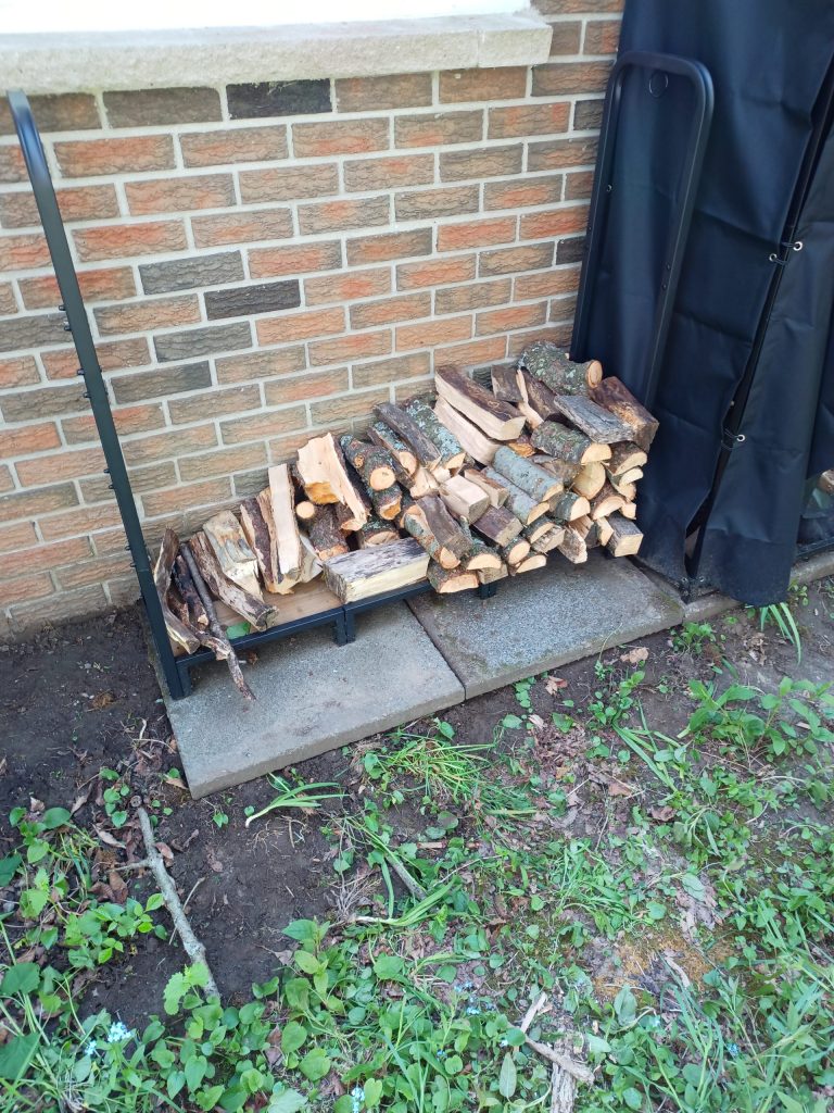 A black steel rack about 120cm wide and 120cm high. The rack is about 15% filled with two rows of wood, with shorter pieces in the front row. Each piece of wood is on average less than 6cm by 6cm when viewed from the end.